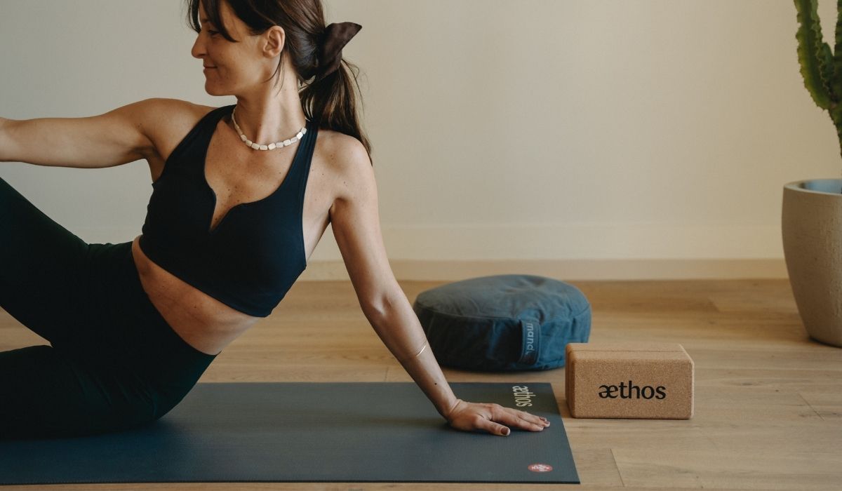 Woman in black workout clothes doing yoga on a mat next to a cushion and a cork yoga block in a minimalist room.