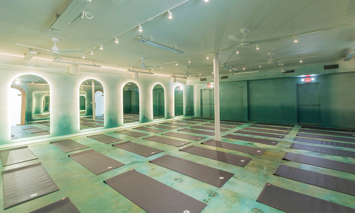 Yoga studio with multiple gray mats neatly arranged on a green floor, arched mirrors with glowing backlights, and ceiling fans.