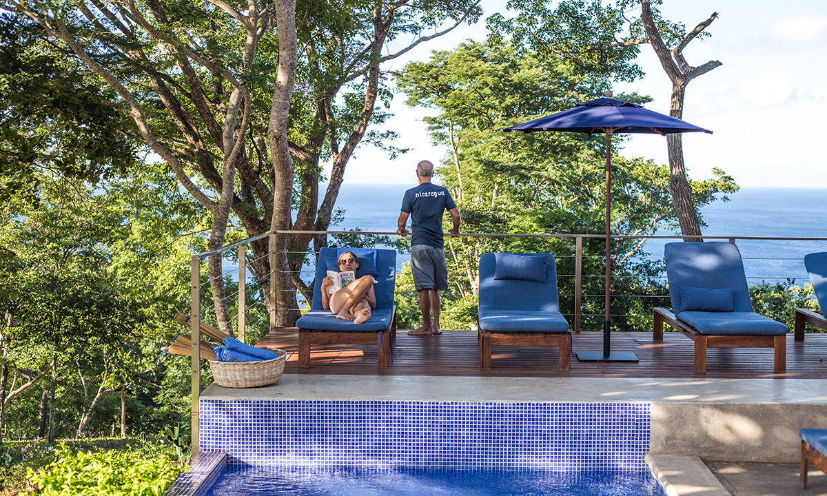 A serene poolside scene with a person lounging on a blue chair, reading, while another stands near the railing. Ocean and trees create a peaceful, sunny backdrop.