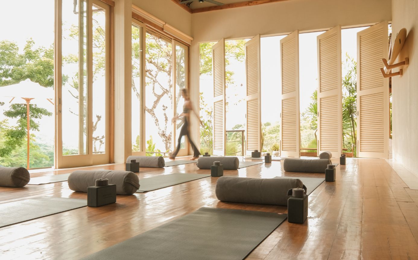 Bright yoga studio with large windows, yoga mats, bolsters, and a person walking, surrounded by lush greenery outside.