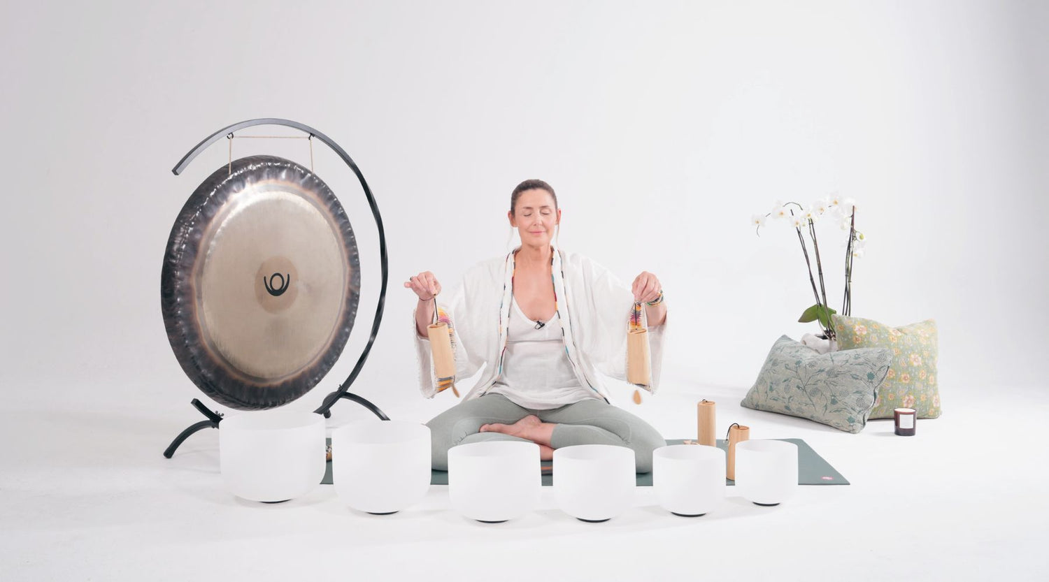 A person sits cross-legged on a yoga mat, holding instruments in each hand, surrounded by singing bowls, a gong, and decorative cushions.