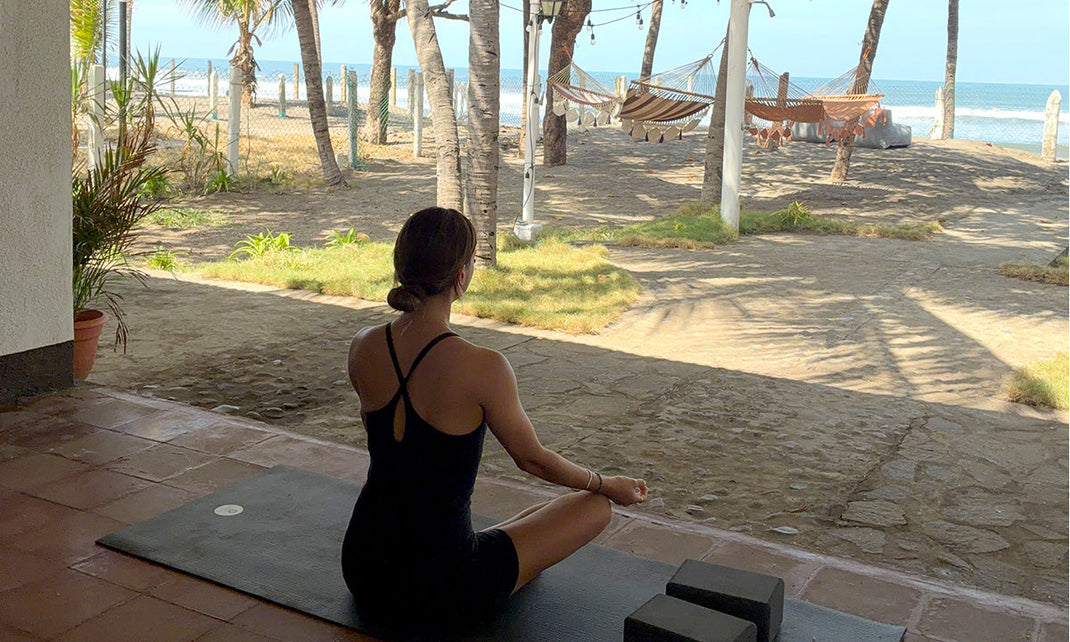 Woman meditating on a yoga mat facing palm trees, hammocks, and the ocean on a sunny day.