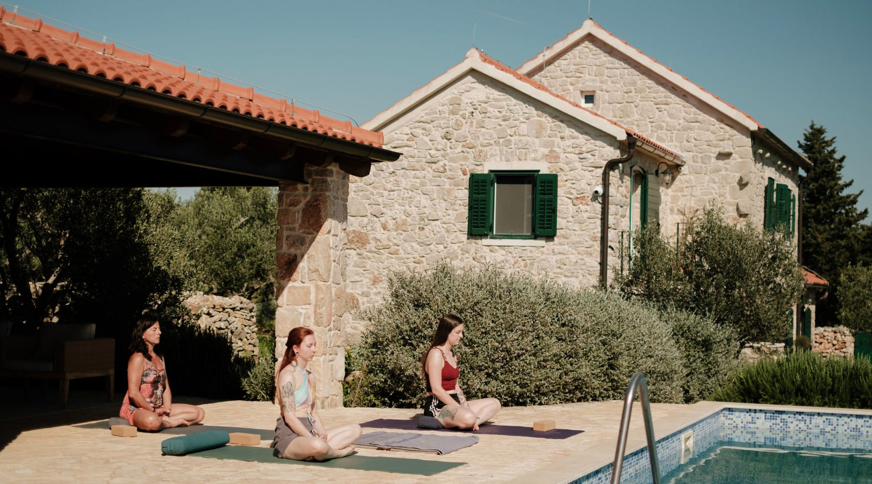 Three women practice yoga on colorful mats by a stone house, surrounded by olive trees and a serene swimming pool under a clear blue sky.