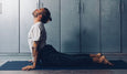 A person performs the Cobra pose on a yoga mat in a minimalist indoor space with gray walls and cabinets.