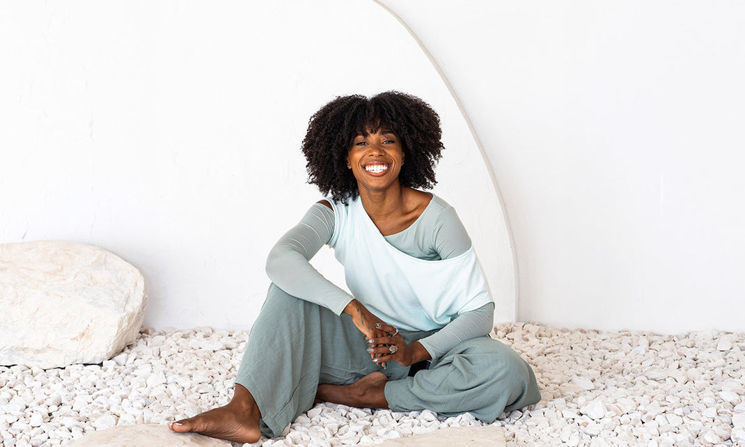 Smiling woman with curly hair in relaxed clothing sitting barefoot on white pebbles against a white wall.