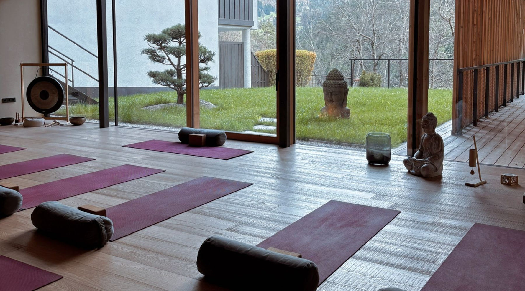 A serene yoga studio with dark red yoga mats, round bolsters, and large windows showing a green garden and a stone Buddha statue.