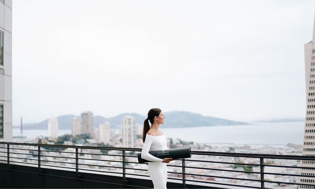 Woman in white outfit holding a yoga mat stands on a terrace overlooking a cityscape with water in the background.