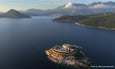 Aerial view of a circular fortress on a rocky islet surrounded by calm blue sea with mountains and scattered clouds in the background.