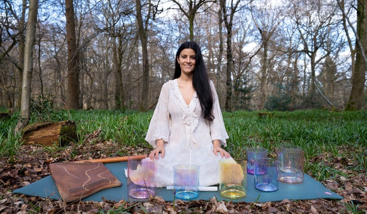 Person in a white dress sits on a mat in a forest, surrounded by colorful crystal singing bowls and a wooden percussion instrument.