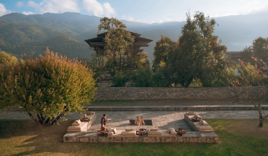 Outdoor seating area with cushions around a central fire pit, surrounded by trees and mountains in the background under a partly cloudy sky.