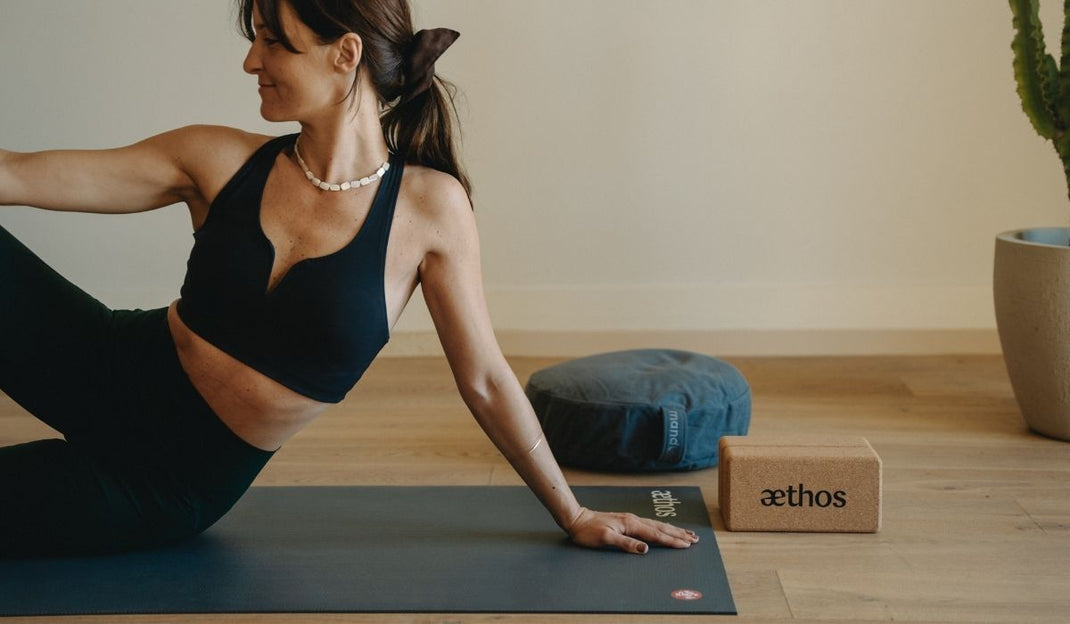 Woman in black workout clothes doing yoga on a mat next to a cushion and a cork yoga block in a minimalist room.