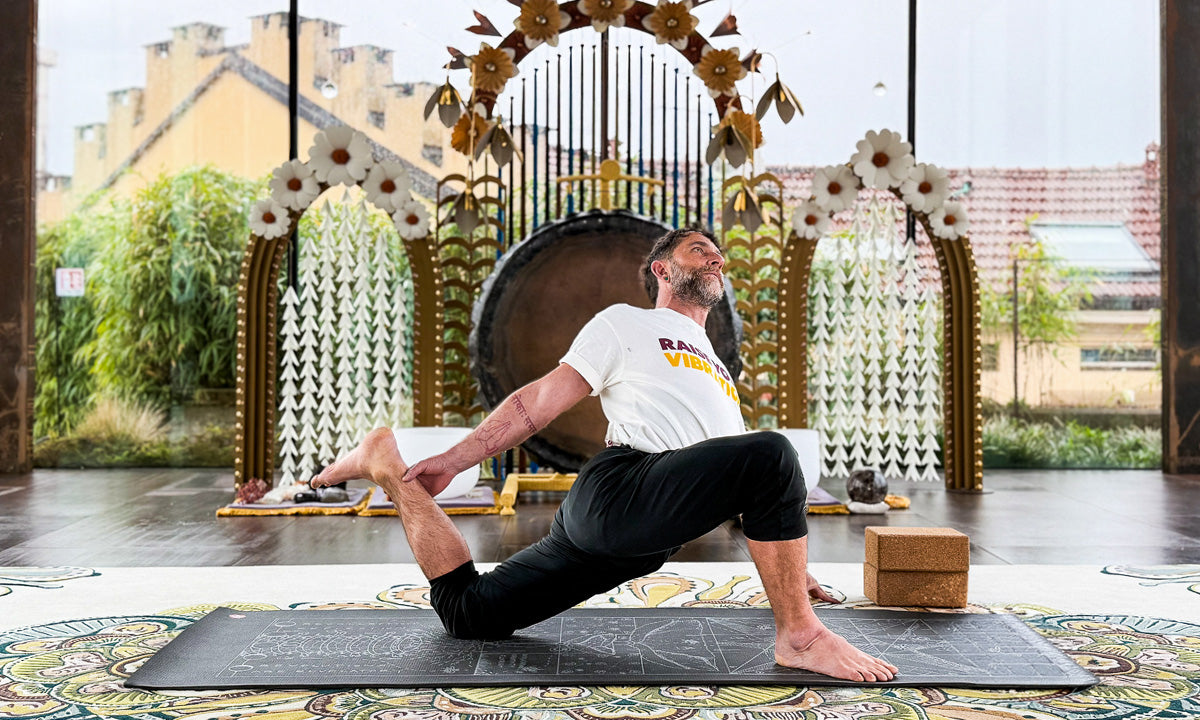 Person in a white shirt and black pants performing a yoga stretch on a mat indoors with decorative arches and a gong in the background.