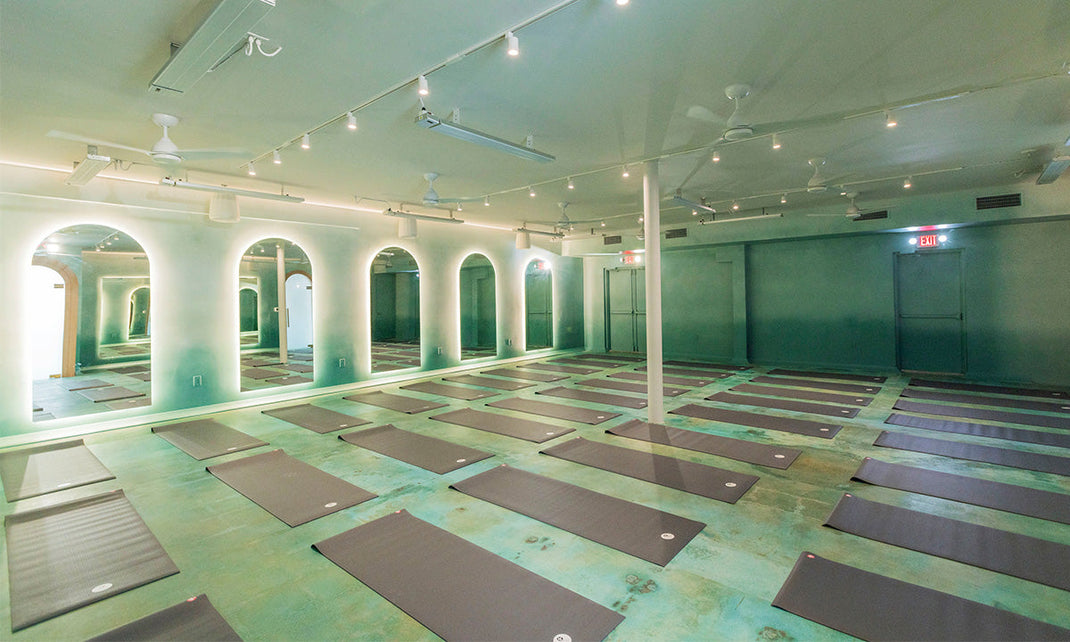 Yoga studio with multiple gray mats neatly arranged on a green floor, arched mirrors with glowing backlights, and ceiling fans.