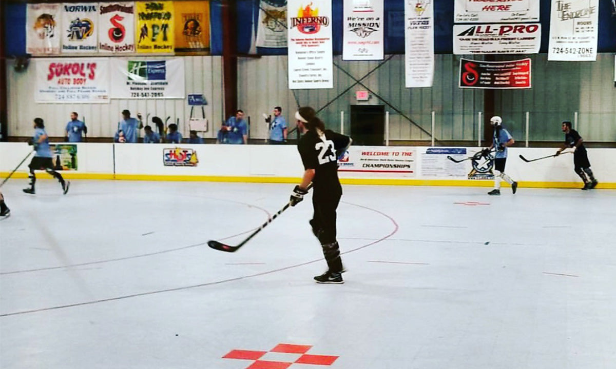 Roller hockey players in action on an indoor rink with banners hanging above and spectators behind the boards.
