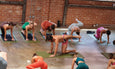 Group yoga class practicing a balance pose on mats in a spacious studio with brick walls and concrete floor.