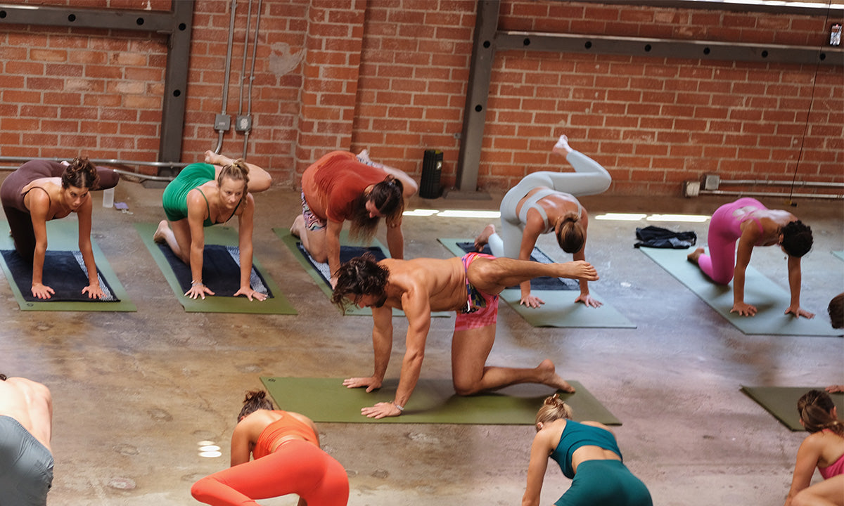 Group yoga class practicing a balance pose on mats in a spacious studio with brick walls and concrete floor.