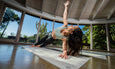 A person performs a side plank yoga pose on a mat, surrounded by large windows offering a view of lush greenery.