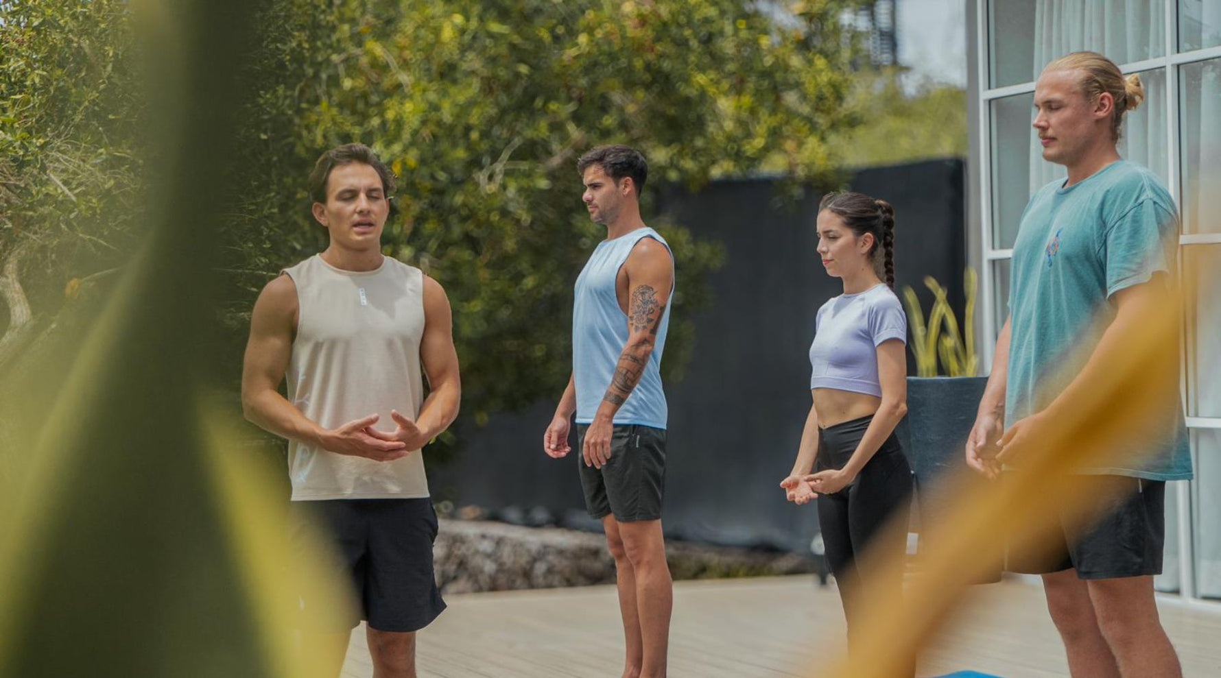 A group of four individuals stand in a yoga pose outdoors, surrounded by greenery and natural sunlight.