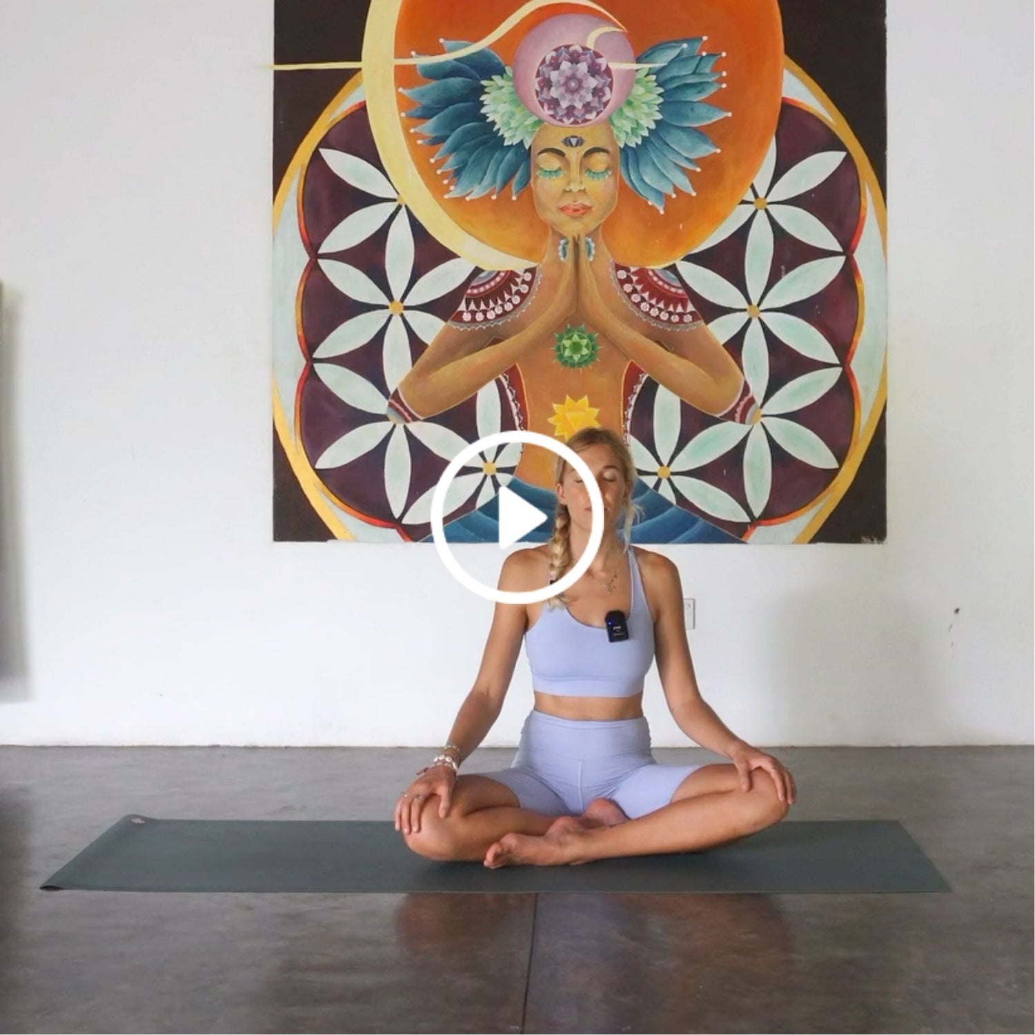 Woman in a purple outfit meditating cross-legged on a mat, with a colorful spiritual mural behind her on a white wall.
