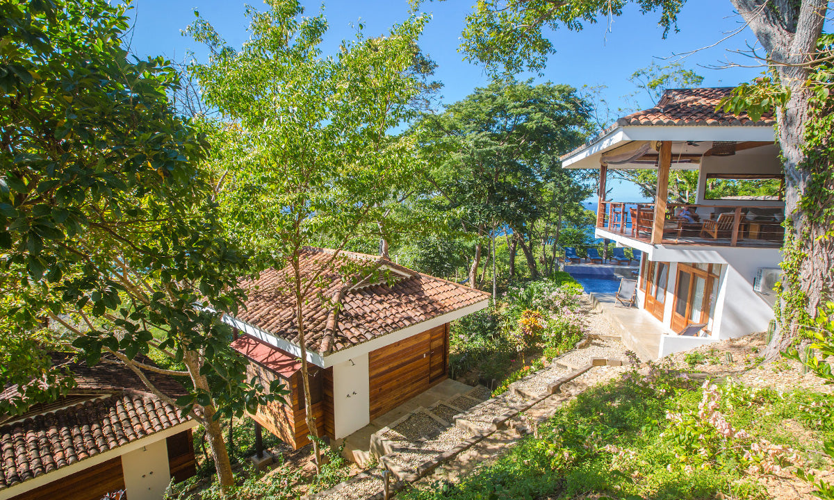A scenic view of a tropical villa with wooden cabins, surrounded by lush greenery and a pool with ocean views.