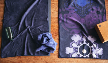 Two hot yoga towels in deep purple tones laid out on a wooden surface, with a green water bottle and a blue towel nearby.