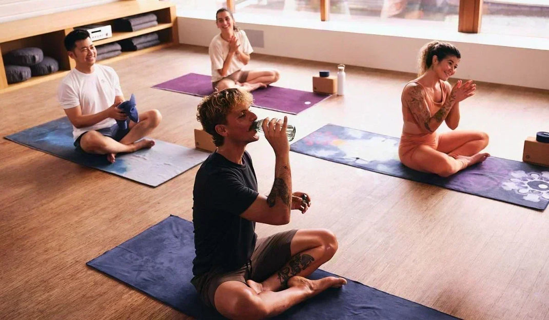 People sit cross-legged on yoga mats in a bright studio, drinking water and preparing for a class.