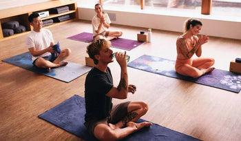 People sit cross-legged on yoga mats in a bright studio, drinking water and preparing for a class.