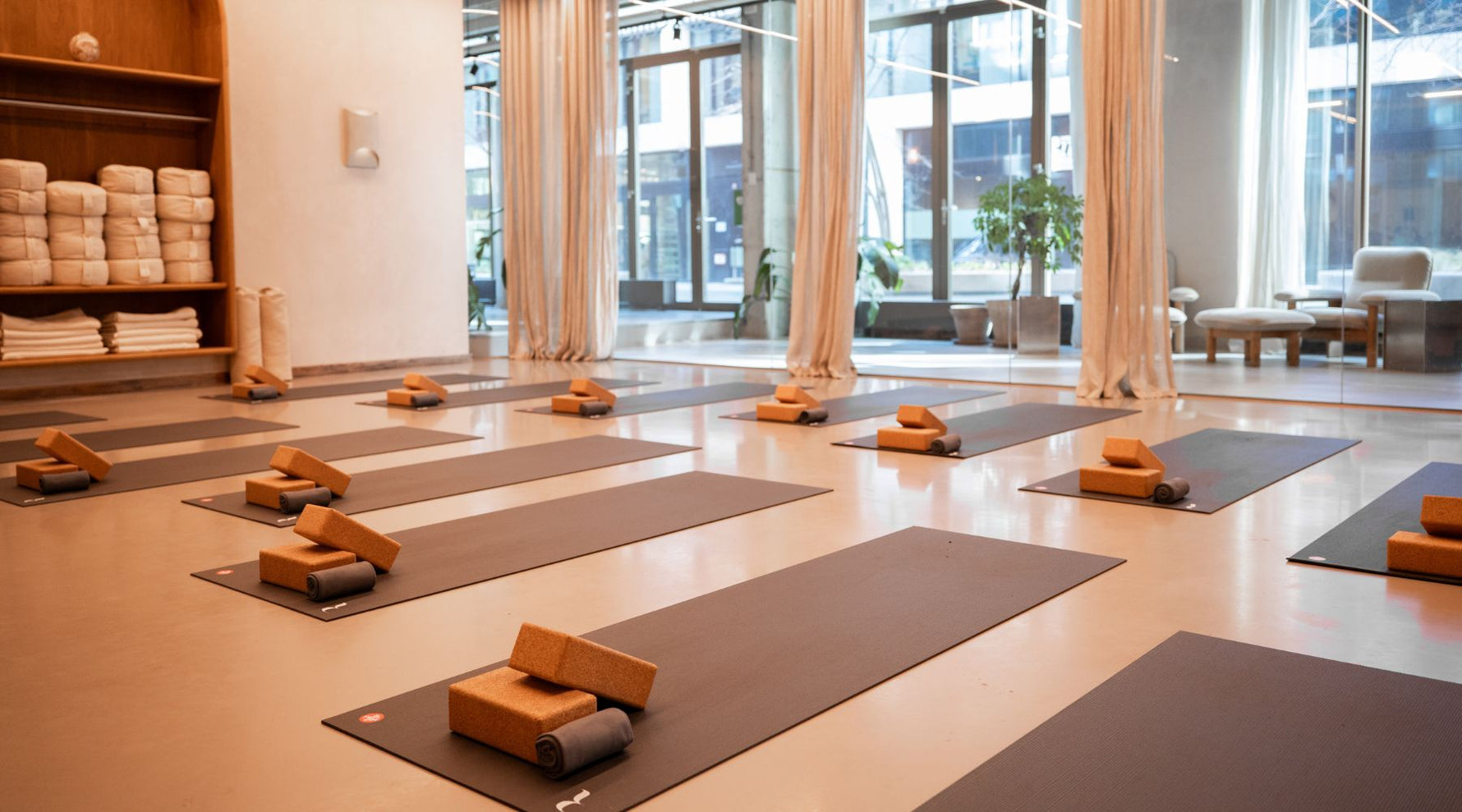A serene yoga studio with neatly arranged mats, cork blocks, and rolled towels, framed by large windows and soft curtains.