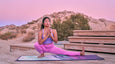 A woman in a purple outfit performs a yoga pose on a colorful mat, set against a desert backdrop and a pastel sunset sky.