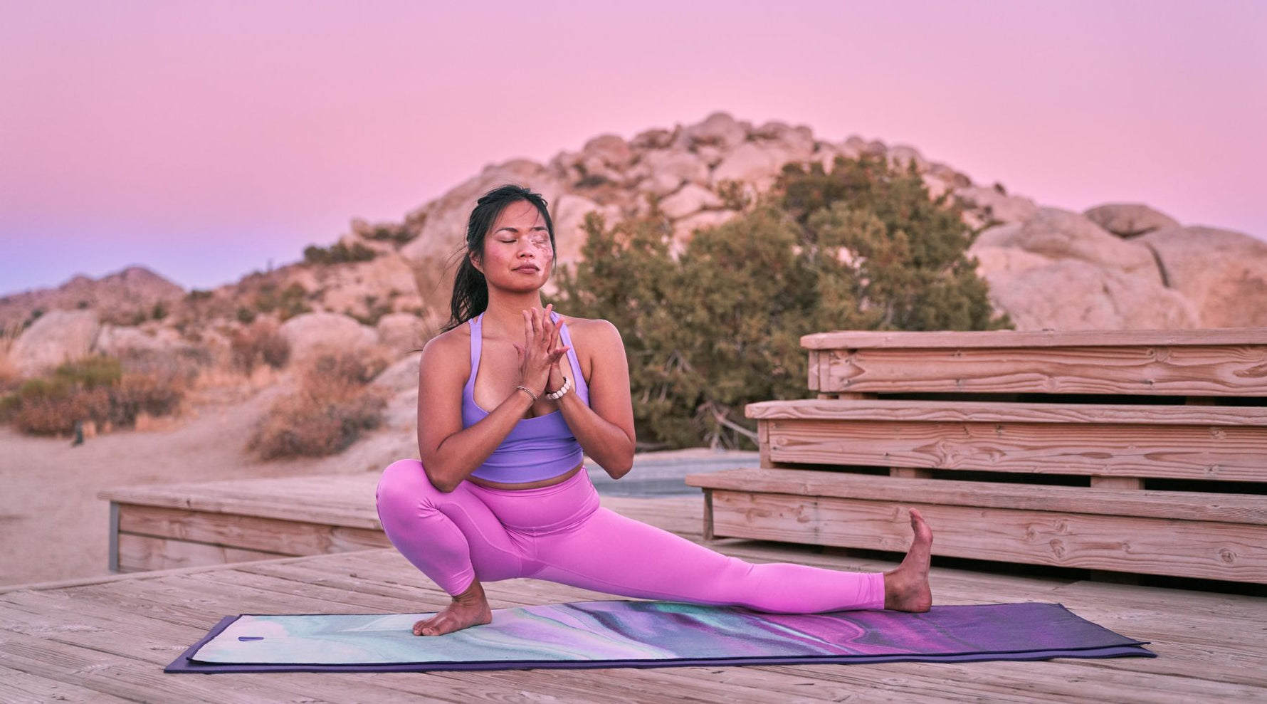 A woman in a purple outfit performs a yoga pose on a colorful mat, set against a desert backdrop and a pastel sunset sky.