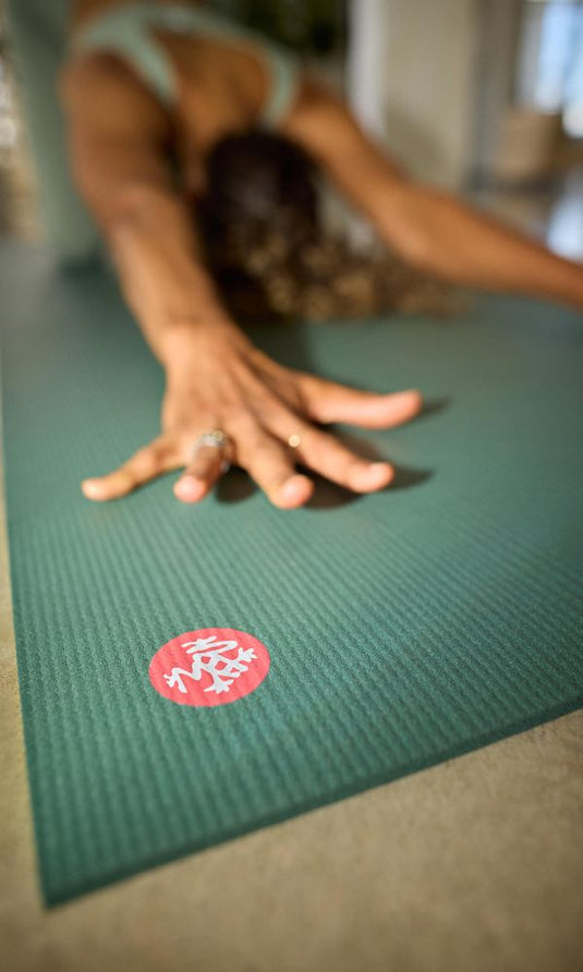 Person reaching forward in a yoga stretch on a textured green Manduka yoga mat with a red circular logo indoors