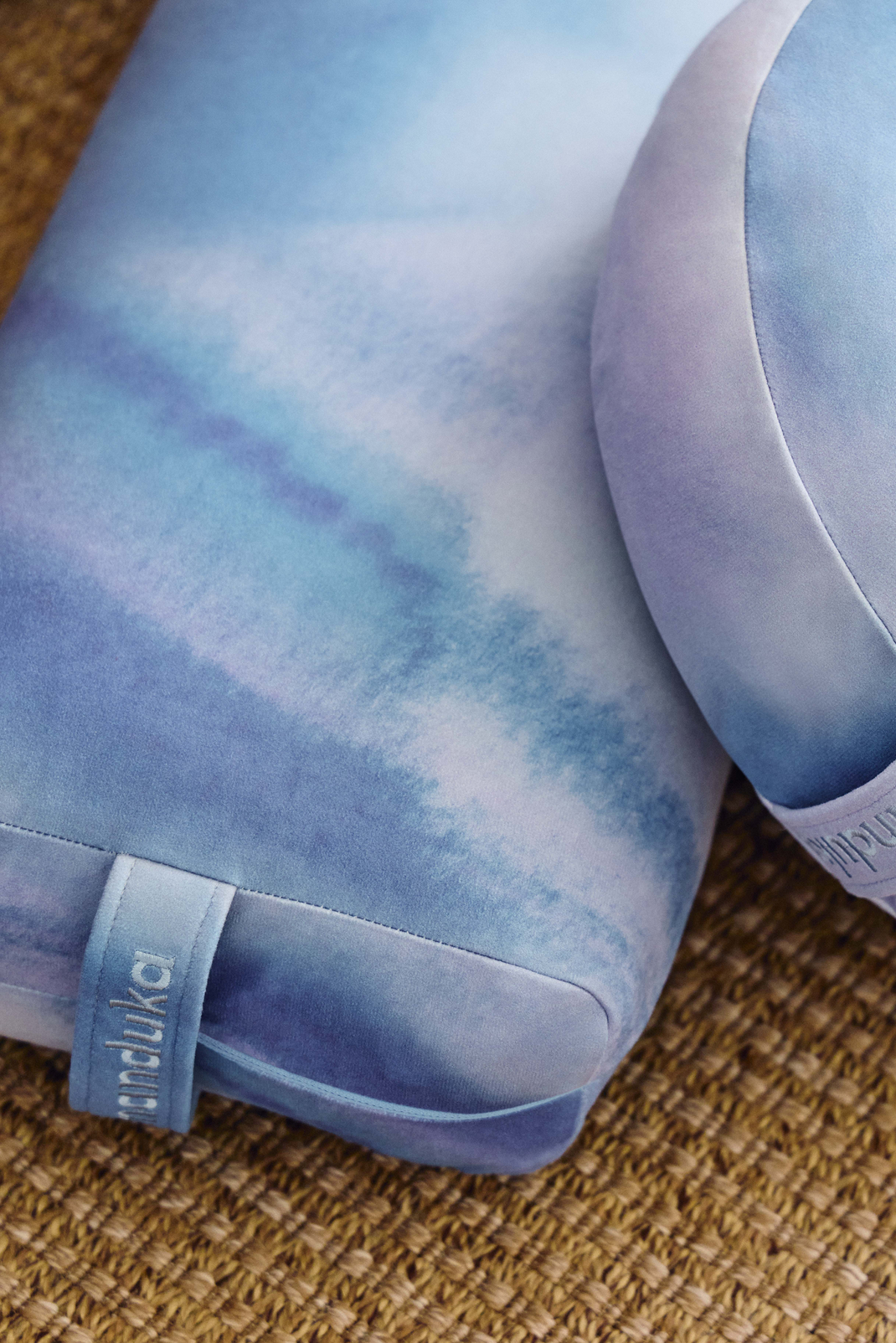 Close up of a blue and lavender marbled Manduka yoga mat with a branded fabric tag and rolled edge resting on a woven brown surface