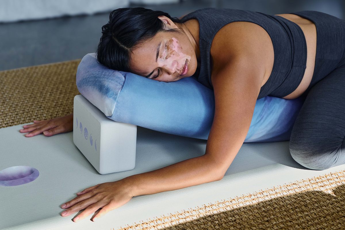 Woman resting in a supported yoga pose on a Manduka yoga mat with a blue bolster and white yoga block