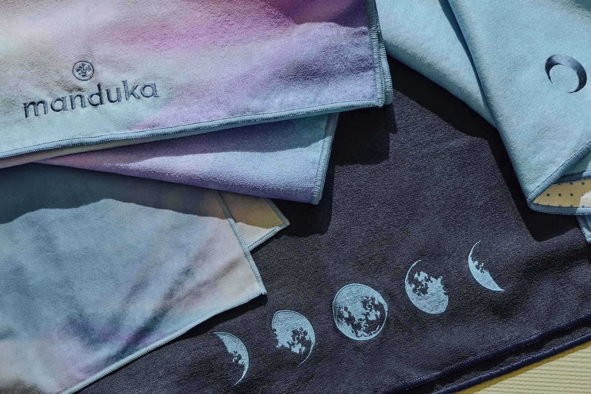 Manduka branded yoga towels in pastel tie dye colors layered over black fabric with white lunar phase designs