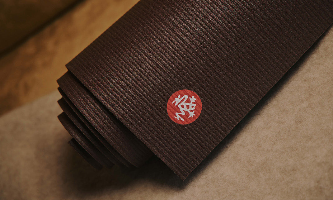 Stack of dark brown Manduka yoga mats with textured ridges and a red circular logo resting on a neutral surface