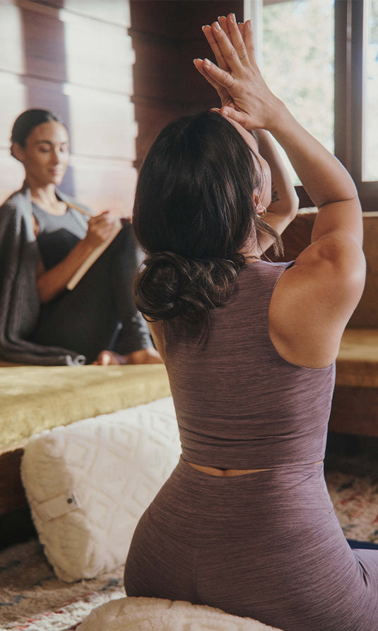 Woman practicing seated yoga with arms raised in a sunlit room, while another person sits in the background near a window.