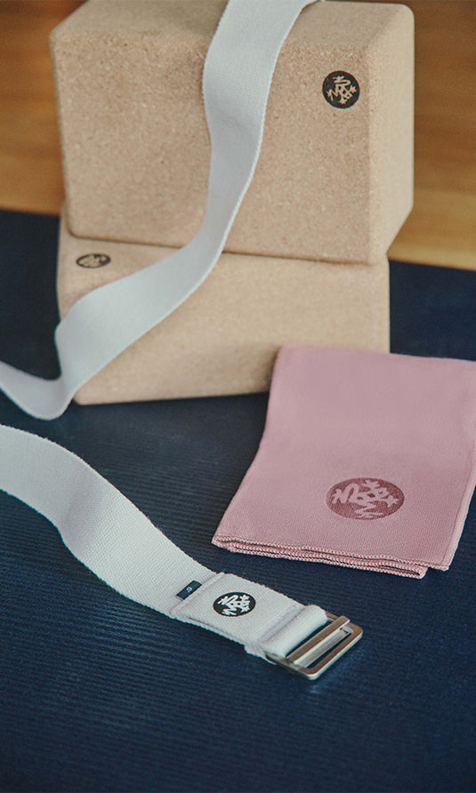 Manduka yoga strap with a metal buckle beside beige cork yoga blocks and a folded pink yoga towel on a dark blue yoga mat