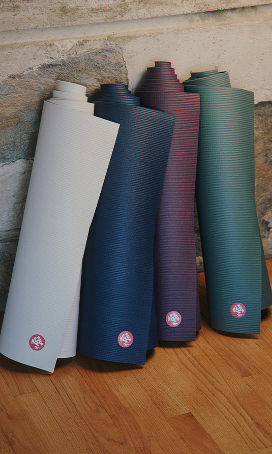 Manduka yoga mats in white, navy, burgundy, and green arranged against a rustic wall on a wooden floor, showing their textured surfaces and rolled ends