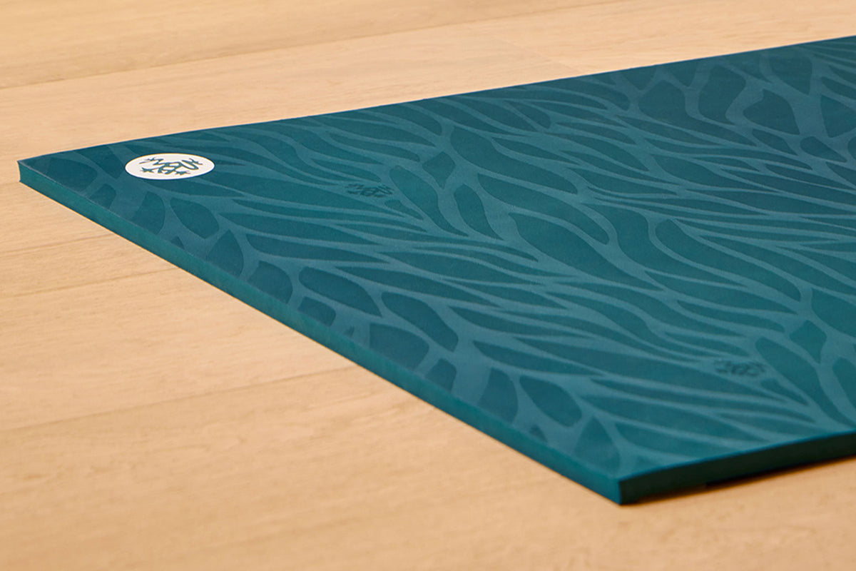 Teal yoga mat with an organic leaf pattern and circular logo resting on a light wood floor
