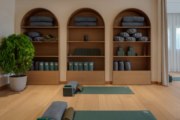 Calm yoga studio with Manduka yoga mats on a wooden floor, folded blankets and bolsters on built in shelves, and a potted plant by the window