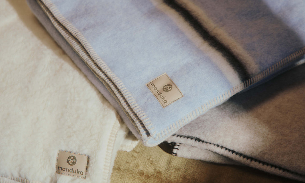 Stack of folded Manduka blankets in soft neutral tones, featuring stitched edges, textured fabric, and visible Manduka labels