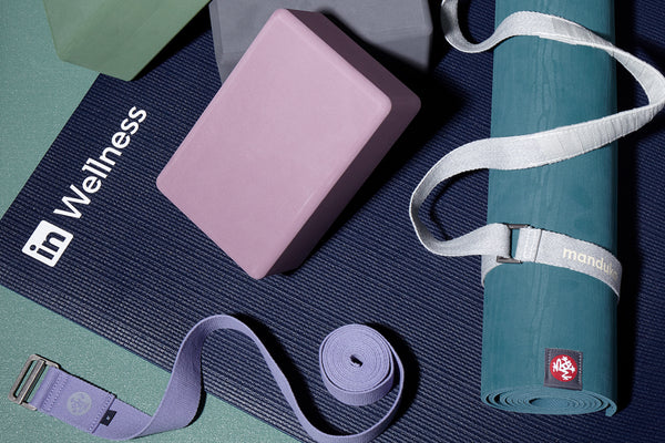 Manduka yoga mat rolled beside pink and green yoga blocks and purple straps on a dark textured mat with Wellness text