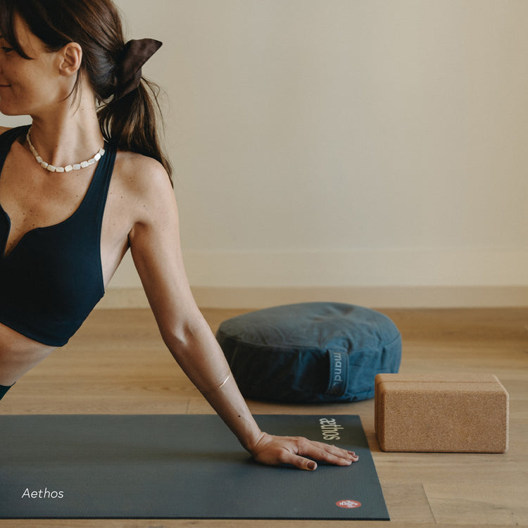 Woman practicing yoga on a Manduka yoga mat beside a cylindrical bolster and cork yoga block in a bright wood floored studio