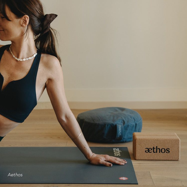 Woman practicing yoga on a dark Manduka yoga mat beside a blue bolster and cork yoga block labeled Aethos in a bright wood floored studio