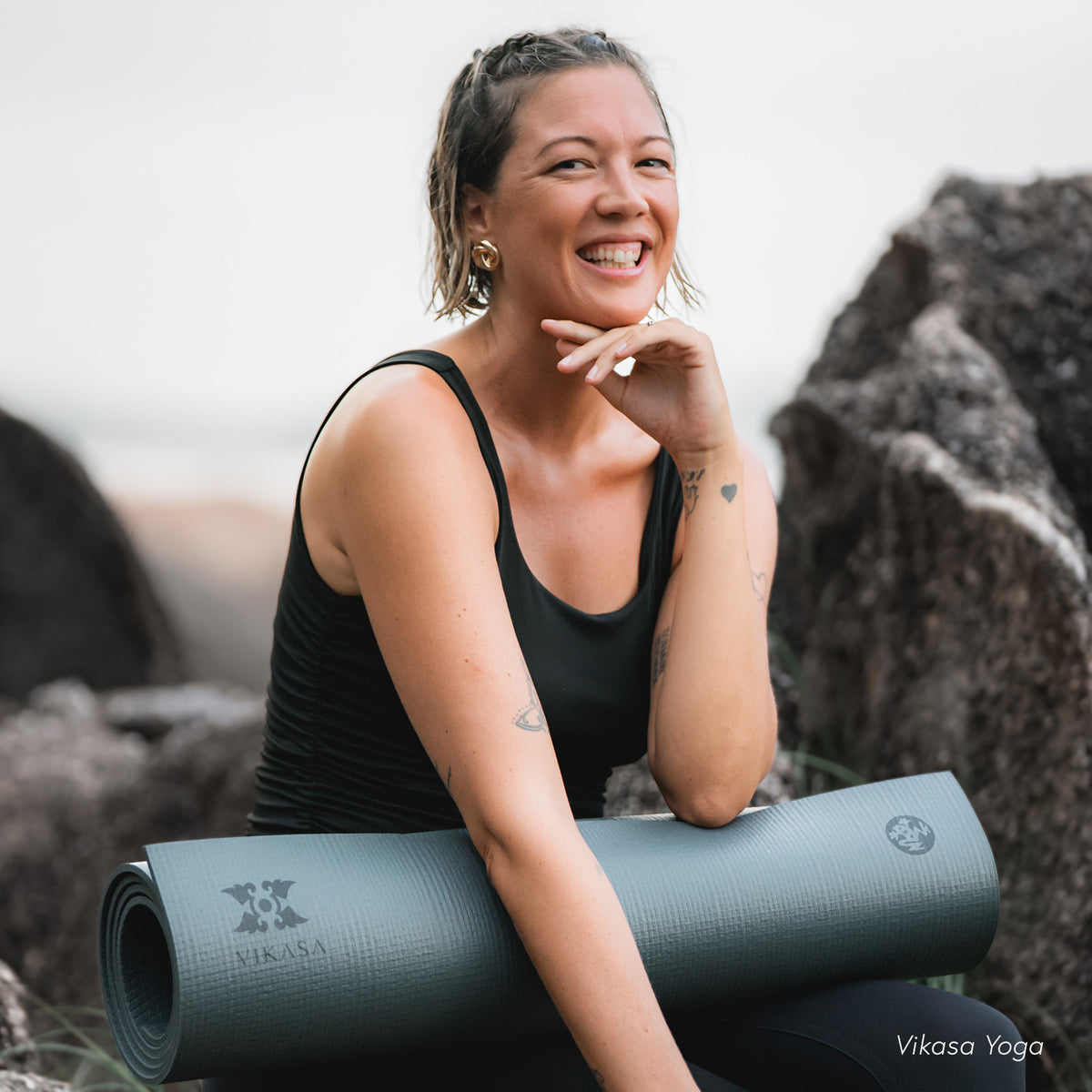 Smiling woman leaning on a rolled Manduka yoga mat outdoors with rocky cliffs in the background and Vikasa Yoga text visible on the image