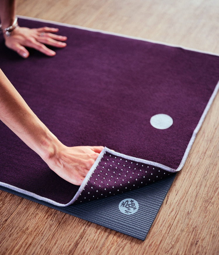 Hands fold a purple Manduka yoga mat to reveal its textured underside on a black yoga mat over a wooden floor