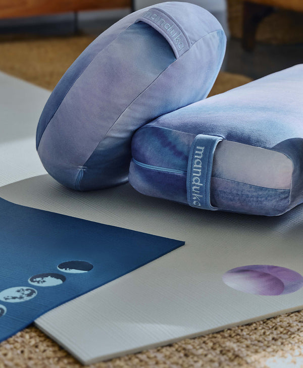 Manduka yoga bolsters in blue and lavender resting on layered yoga mats, with visible Manduka branding and circular design details.