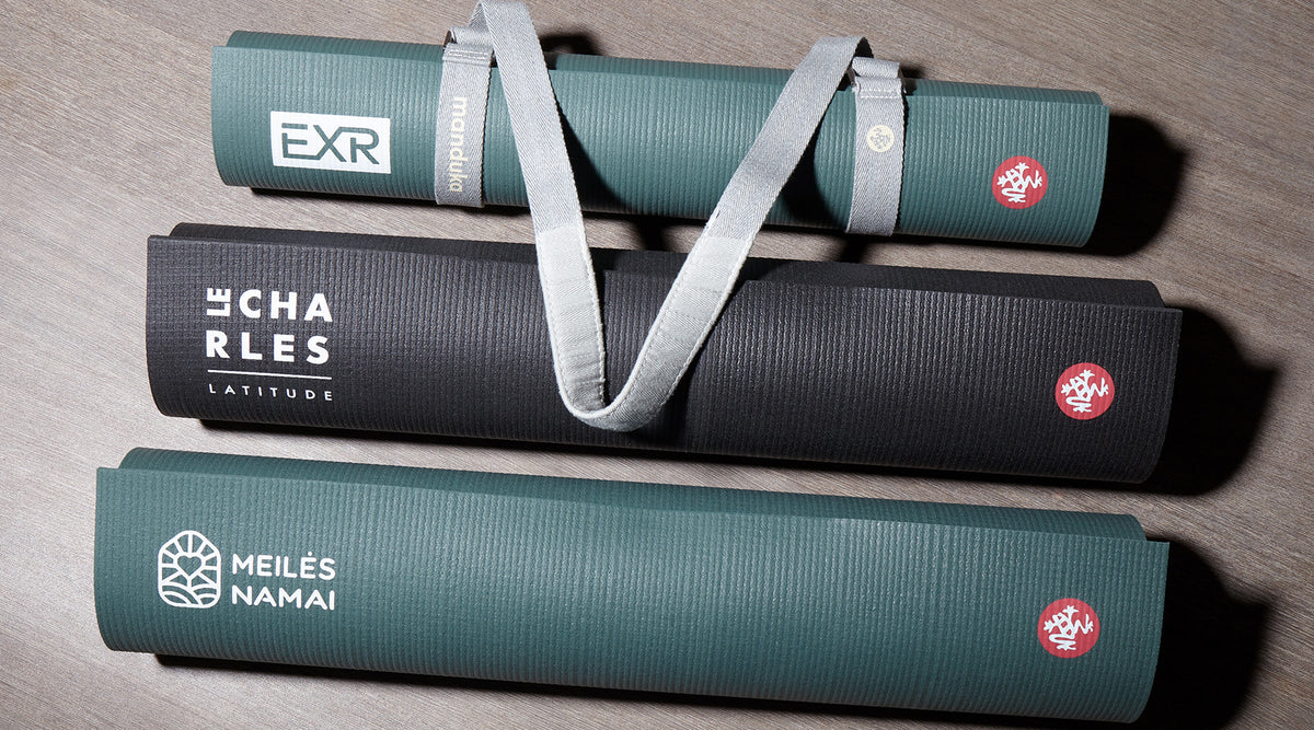 Three rolled Manduka yoga mats in green and black with carrying straps, arranged on a wooden surface and featuring printed logos and text.