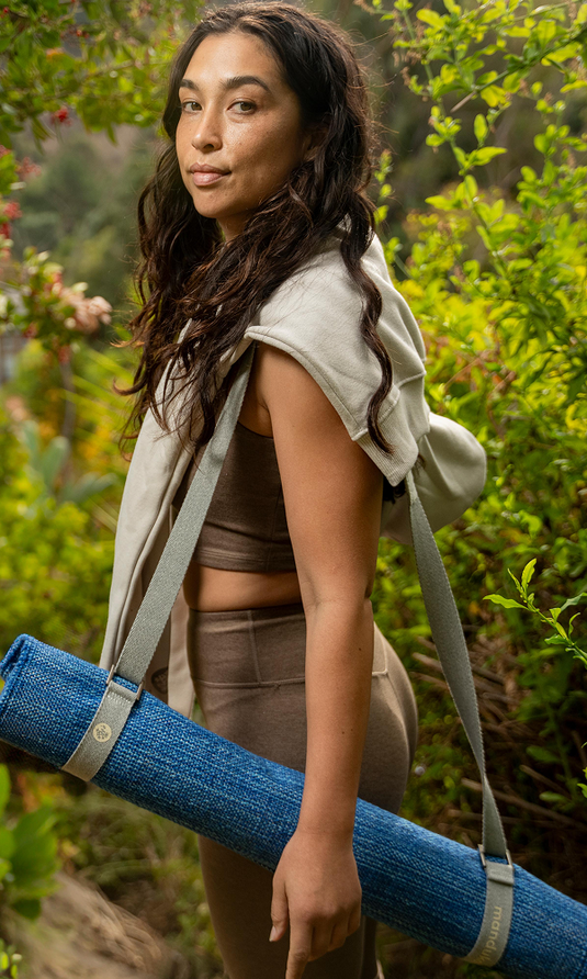 Woman carrying a rolled blue Manduka yoga mat with shoulder straps through a lush garden, wearing neutral athletic clothing and looking toward the camera