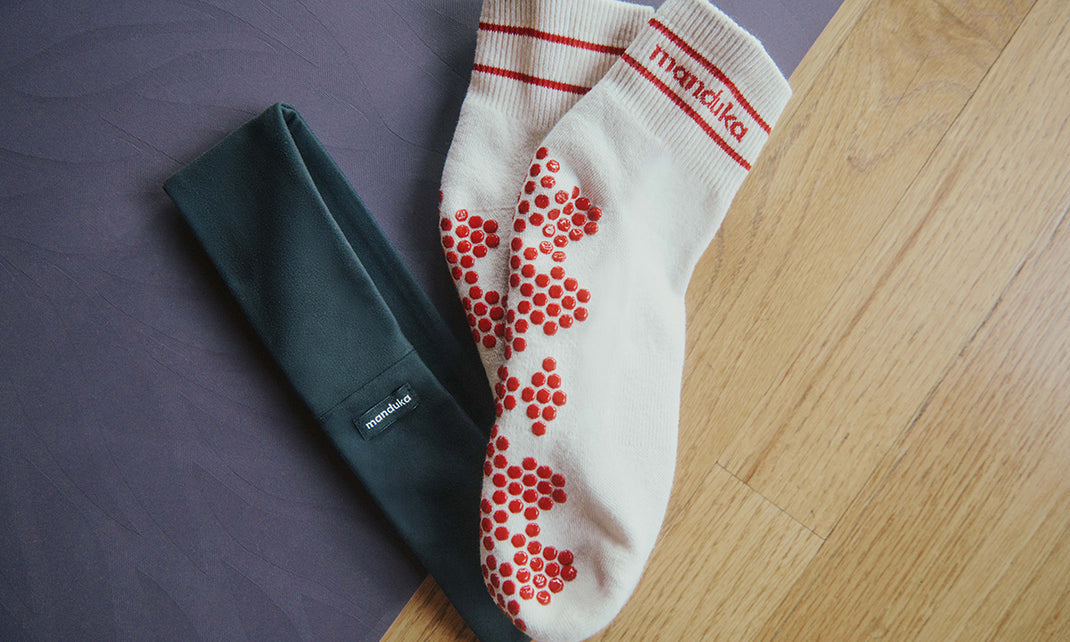 Manduka yoga socks with red grip dots resting on a dark yoga mat beside a light wood floor